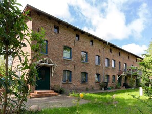 A brick building with a green door and multiple windows, surrounded by greenery and a paved area.