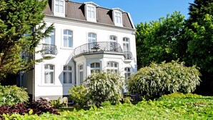 A white building with multiple windows and balconies, surrounded by lush greenery and trees. - 04 Double Room - Villa in the Baltic Sea resort Baabe (Baabe)