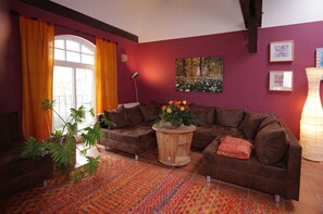 Ein Wohnzimmer mit braunem Sofa, einem Holztisch mit Blumen, einem großen Fenster mit Vorhängen, einem Gemälde an der Wand und einer Lampe.