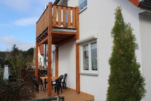 Een huis met twee verdiepingen, met een houten balkon, een eethoek met stoelen en een hoge altijdgroene boom.