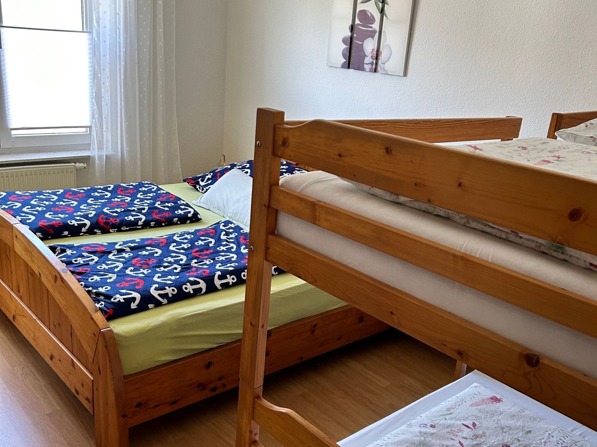 Een houten stapelbed met een onderste bed en een matras op het bovenbed. Er is een raam met dunne gordijnen en een radiator aan de muur.