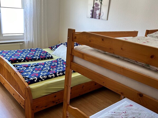 Una cama litera de madera con una cama inferior y un colchón en la litera superior. Hay una ventana con cortinas de gasa y un radiador en la pared.