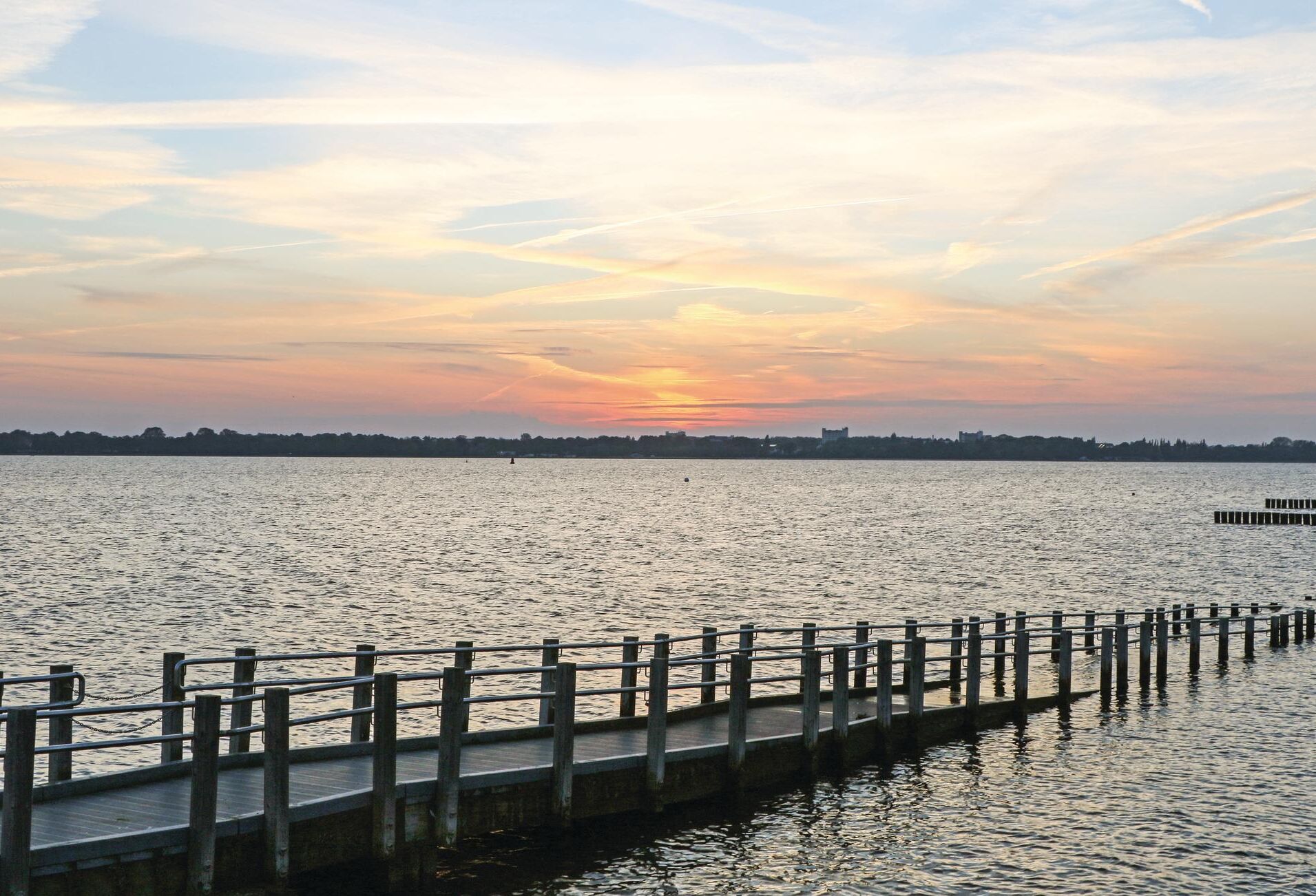 Blick auf das Wasser mit einer Holzsteg, der sich in ruhiges Wasser erstreckt, einer entfernten Skyline und einem Sonnenuntergangshimmel.