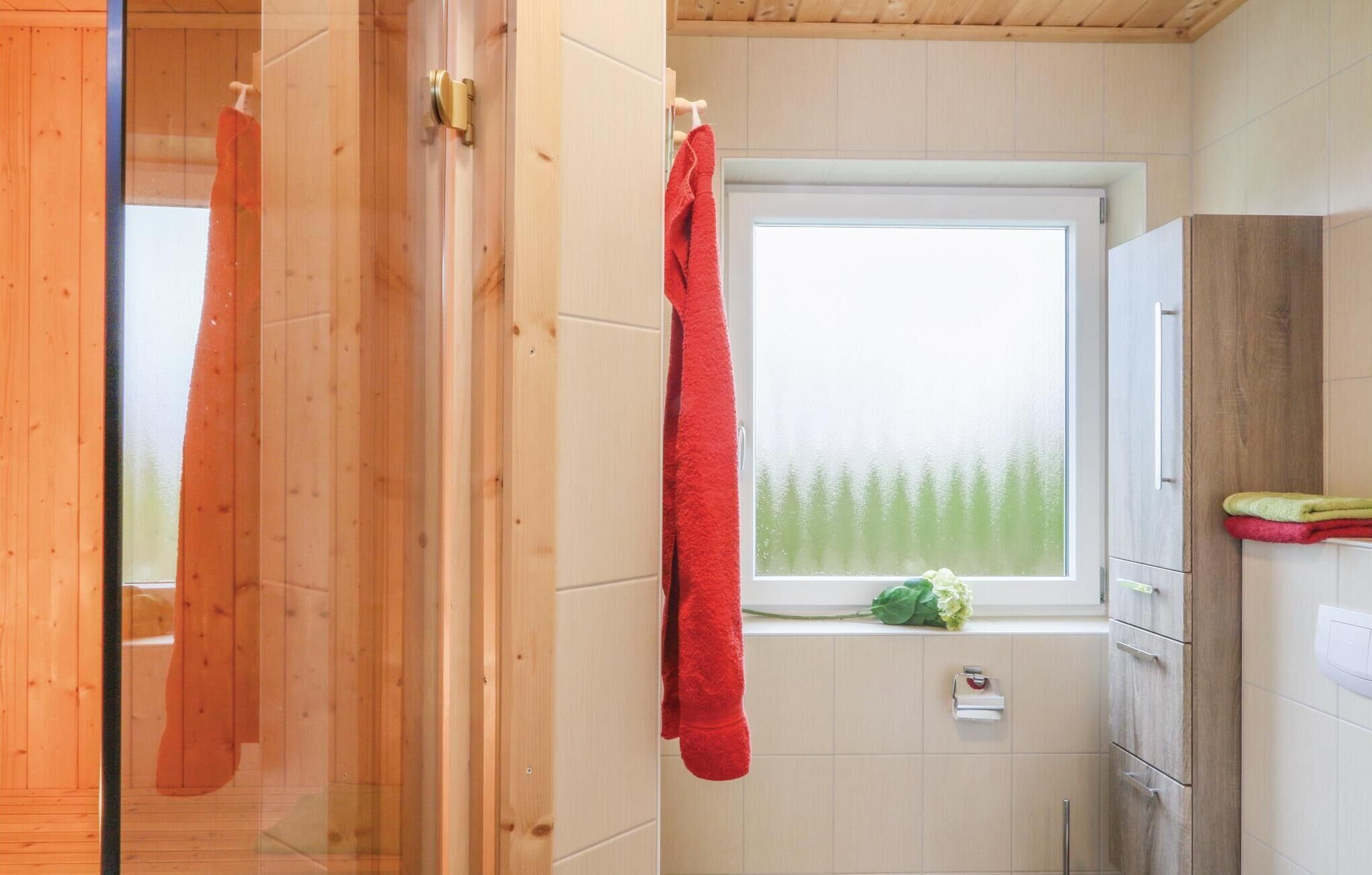 A bathroom with wooden walls, a window with a view of trees, and a red towel hanging on the door.
