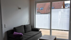 Un salon moderne avec un canapé gris, une petite table en bois et une grande porte-fenêtre coulissante donnant sur une terrasse en bois.
