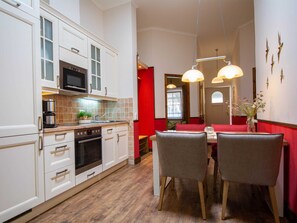 Una cocina moderna con armarios blancos, un microondas y una cafetera. Hay una zona de comedor con mesa y sillas, y una pared de acento roja.