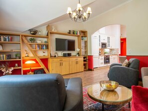 A modern living room with a sofa, a coffee table, and a TV. There is a kitchen in the background with white cabinets and a red accent wall.