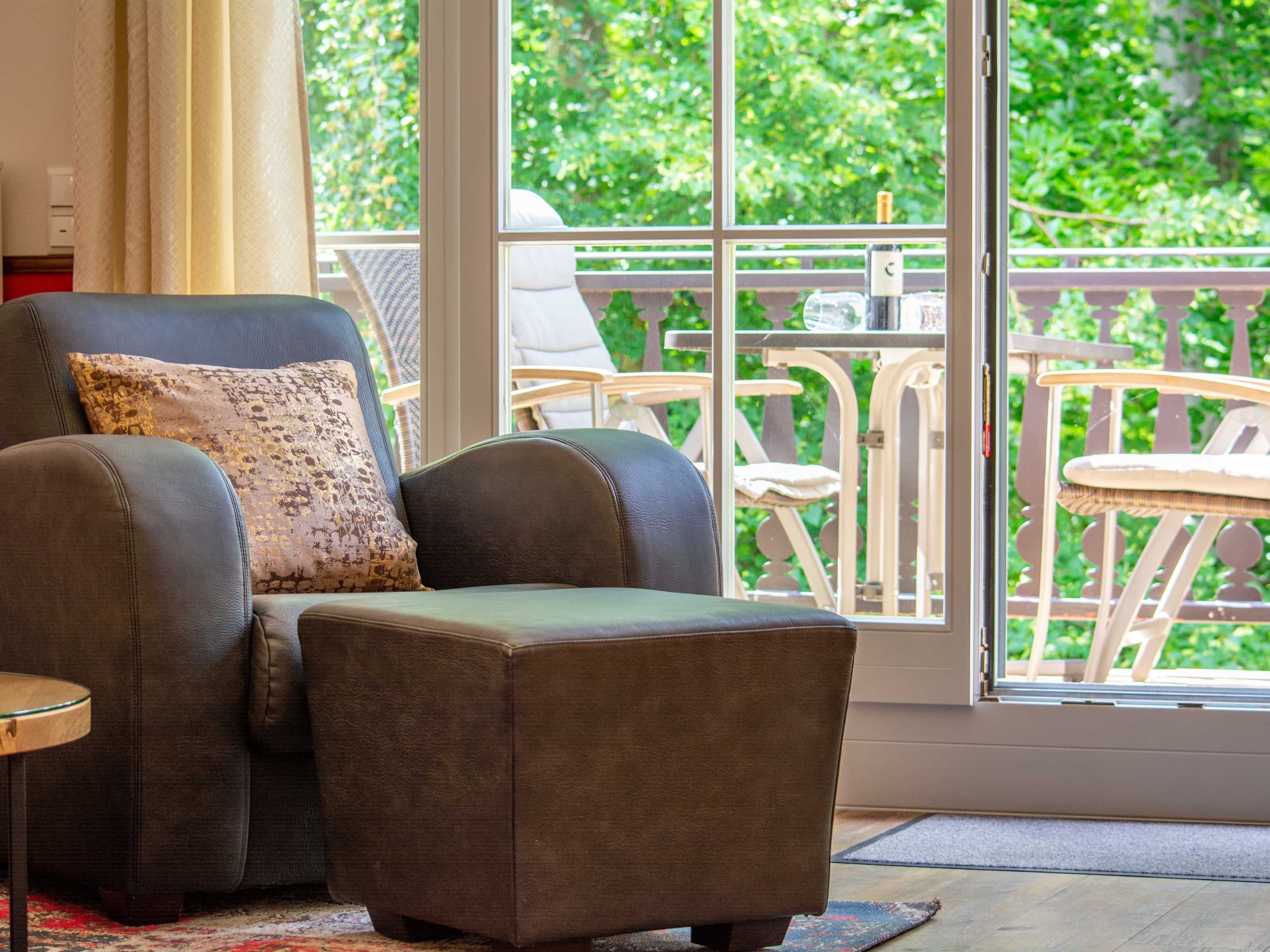 Ein Wohnzimmer mit einer Couch und Hocker, einem kleinen Tisch und Blick auf einen Balkon mit Gartenmöbeln.