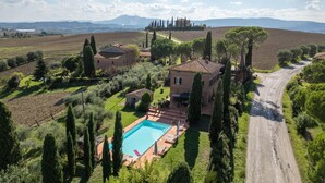 Eine Villa mit Schwimmbad, umgeben von Olivenhainen und Zypressen, mit einer Straße und Hügeln im Hintergrund.