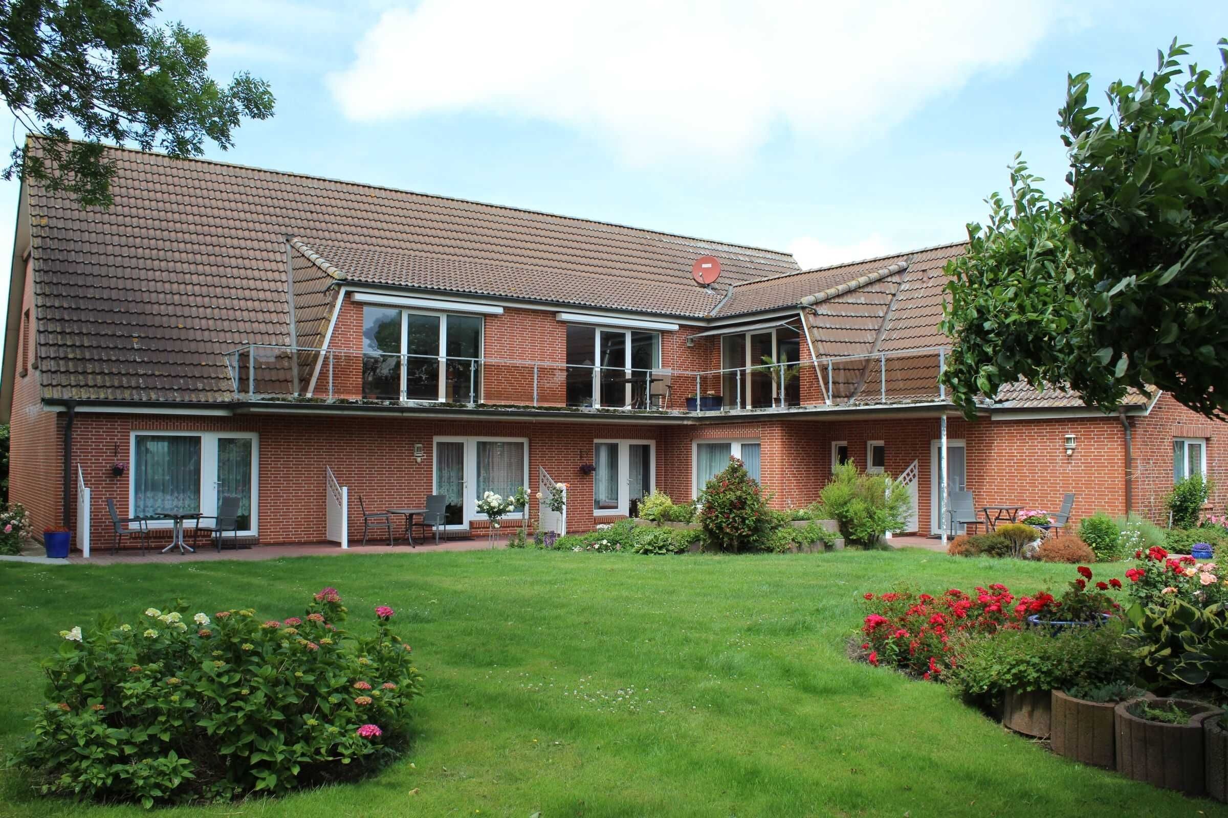 Een herenhuis van baksteen met twee verdiepingen, een overdekte patio, een kleine tuin met diverse planten en bloemen, en een heldere lucht.