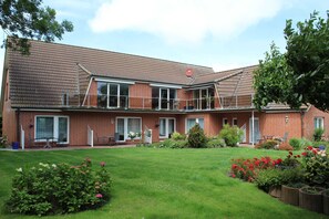 A two-story brick house with a covered patio, a small garden with various plants and flowers, and a clear sky. - Apartment No. 3 - To the little mermaid (Friedrichskoog)