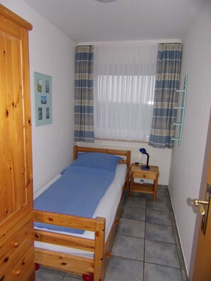 A small, neatly arranged bedroom with a wooden bed, a bedside table with a lamp, a window with curtains, and a wall-mounted shelf. - Apartment No. 3 - To the little mermaid (Friedrichskoog)