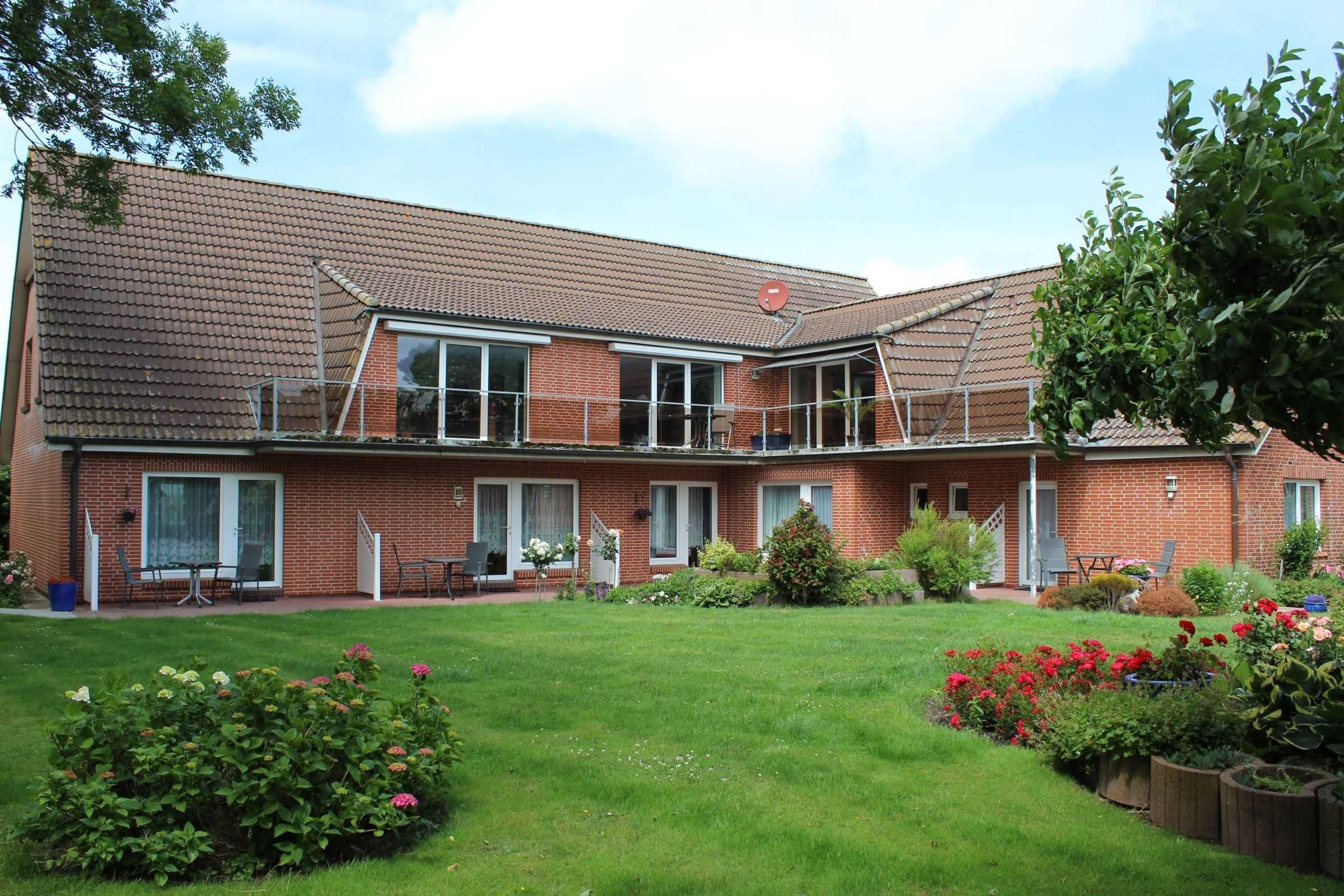 A two-story brick house with a covered patio, a small garden with various plants and flowers, and a clear sky.