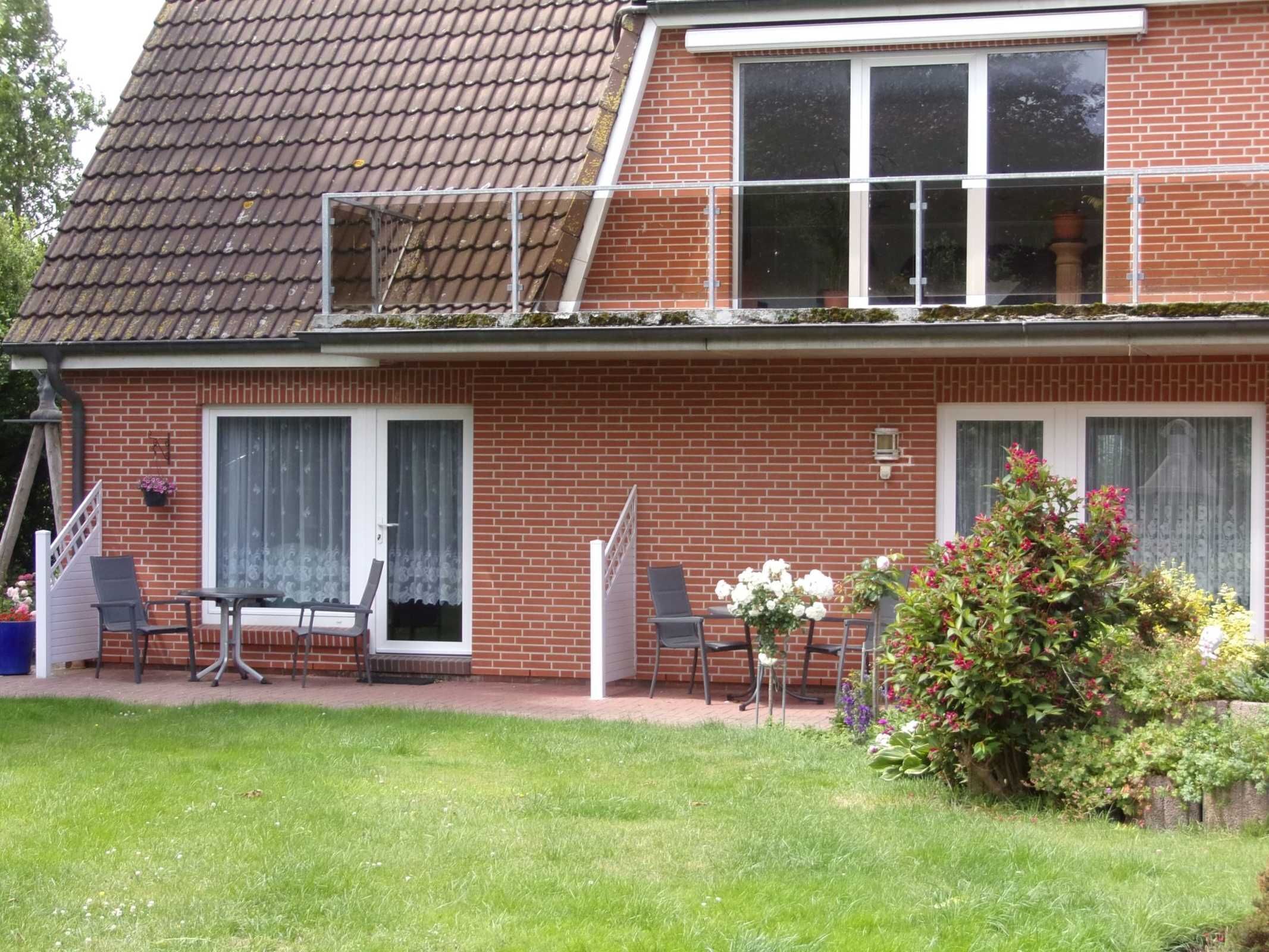 Ein zweistöckiges Backsteinhäuschen mit Ziegeldach, einem Balkon und einem kleinen Gartenbereich mit Stühlen und Blumen.