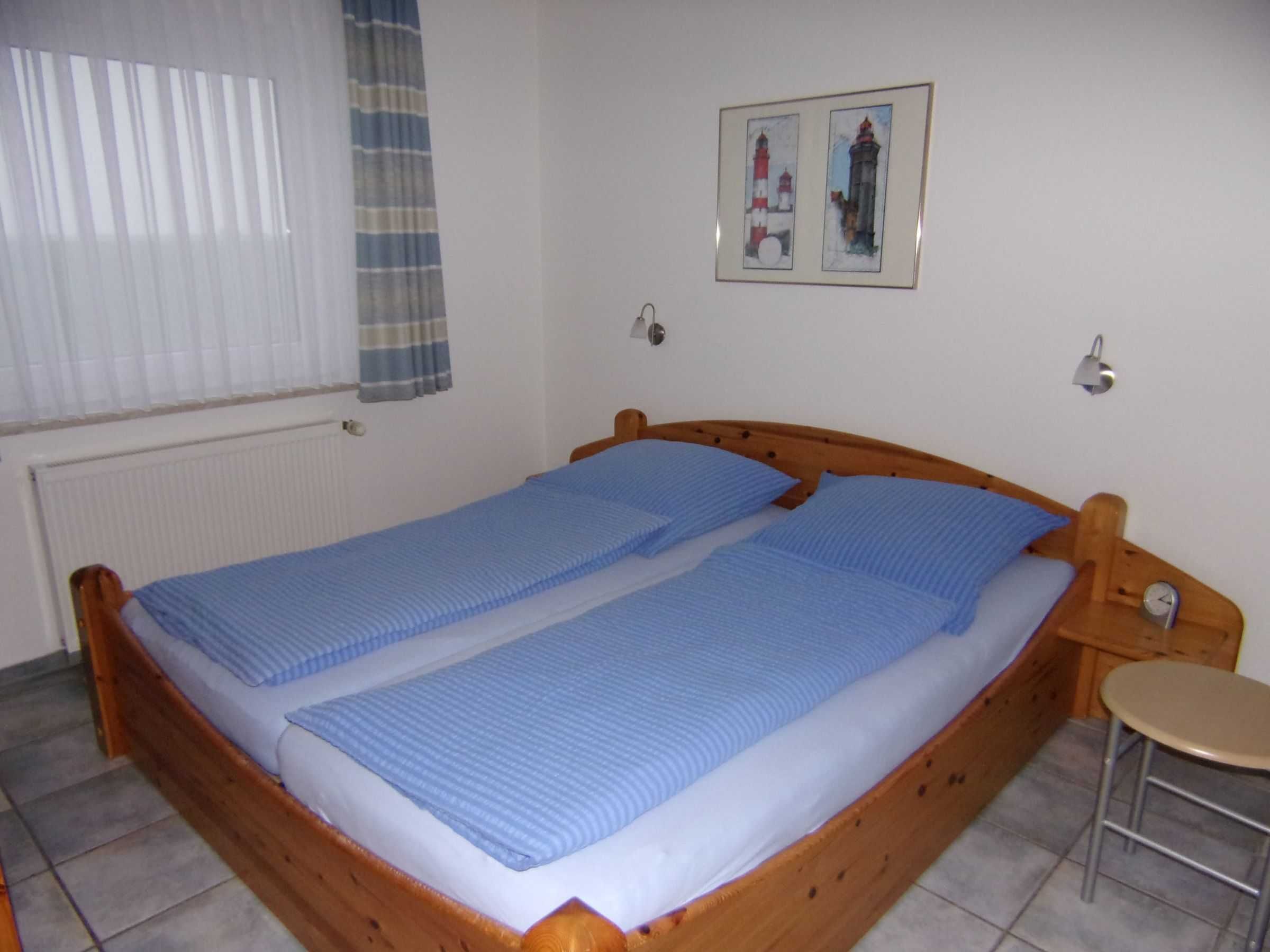 Een eenpersoonskamer met een houten bedframe, een blauwe spreidek en een klein krukje, met een ingelijste foto aan de muur.