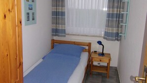 Une petite chambre à coucher, bien rangée, avec un lit en bois, une table de chevet avec une lampe, une fenêtre avec des rideaux et une étagère fixée au mur.