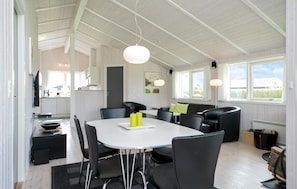 A modern dining area with a white table, black chairs, and a sofa. There are pendant lights, a view of the outdoors, and a kitchen in the background. - Lovely home in Hadsund with sauna (Hadsund)