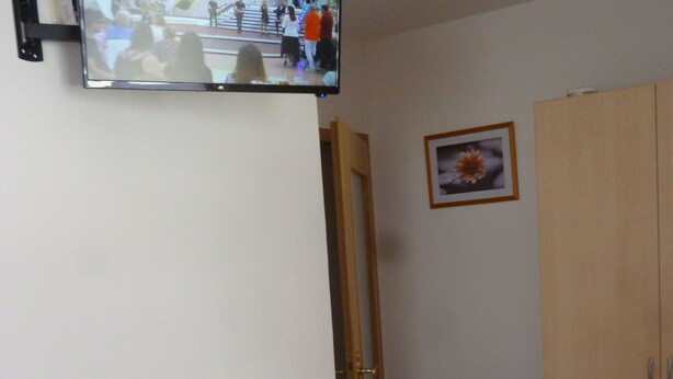 A small room with a wooden table, two stools, a TV mounted on the wall, a wooden wardrobe, and a framed picture on the wall. - Dormitory Room - Small Room (Heiligenstadt i.OFr.)