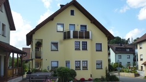 A two-story building with a balcony, multiple windows, and a paved courtyard with flower beds. - Dormitory Room - Small Room (Heiligenstadt i.OFr.)