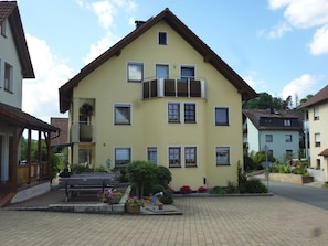 A two-story building with a balcony, multiple windows, and a paved courtyard with flower beds. - Dormitory Room - Small Room (Heiligenstadt i.OFr.)