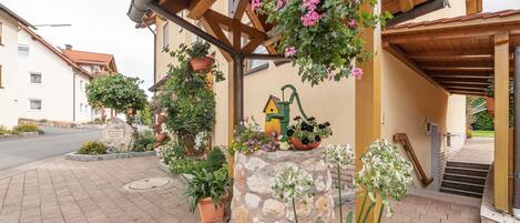 Un pilier en pierre surmonté d’un jardinier en forme de maison, une pergola en bois ornée de plantes suspendues et un chemin pavé menant à l’entrée d’un bâtiment.