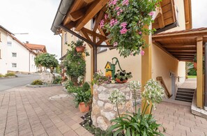 A stone pillar with a house-shaped planter, a wooden pergola with hanging flowers, and a paved walkway leading to a building entrance. - Dormitory Room - Small Room (Heiligenstadt i.OFr.)