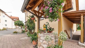 A stone pillar with a house-shaped planter, a wooden pergola with hanging flowers, and a paved walkway leading to a building entrance. - Dormitory Room - Small Room (Heiligenstadt i.OFr.)