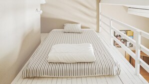 A small, single-bed room with a striped bedspread and pillows, a white railing, and a view of a dining area through the window. - Amazing home in Rochefort (Rochefort)