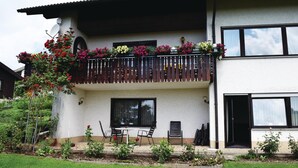 Een gebouw met twee verdiepingen, voorzien van een balkon versierd met kleurrijke bloemen, een klein terras met stoelen, en een tuin met diverse planten.