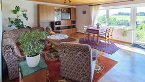 Een woonkamer met een bank, fauteuil, eettafel en stoelen. Er staat een potplant, een televisie en een raam met uitzicht op de omgeving.