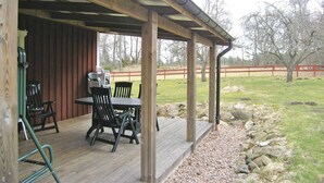 Ein überdachter Außenbereich mit Tisch und Stühlen, einer Holzterrasse und Blick auf einen umzäunten Garten mit Bäumen.