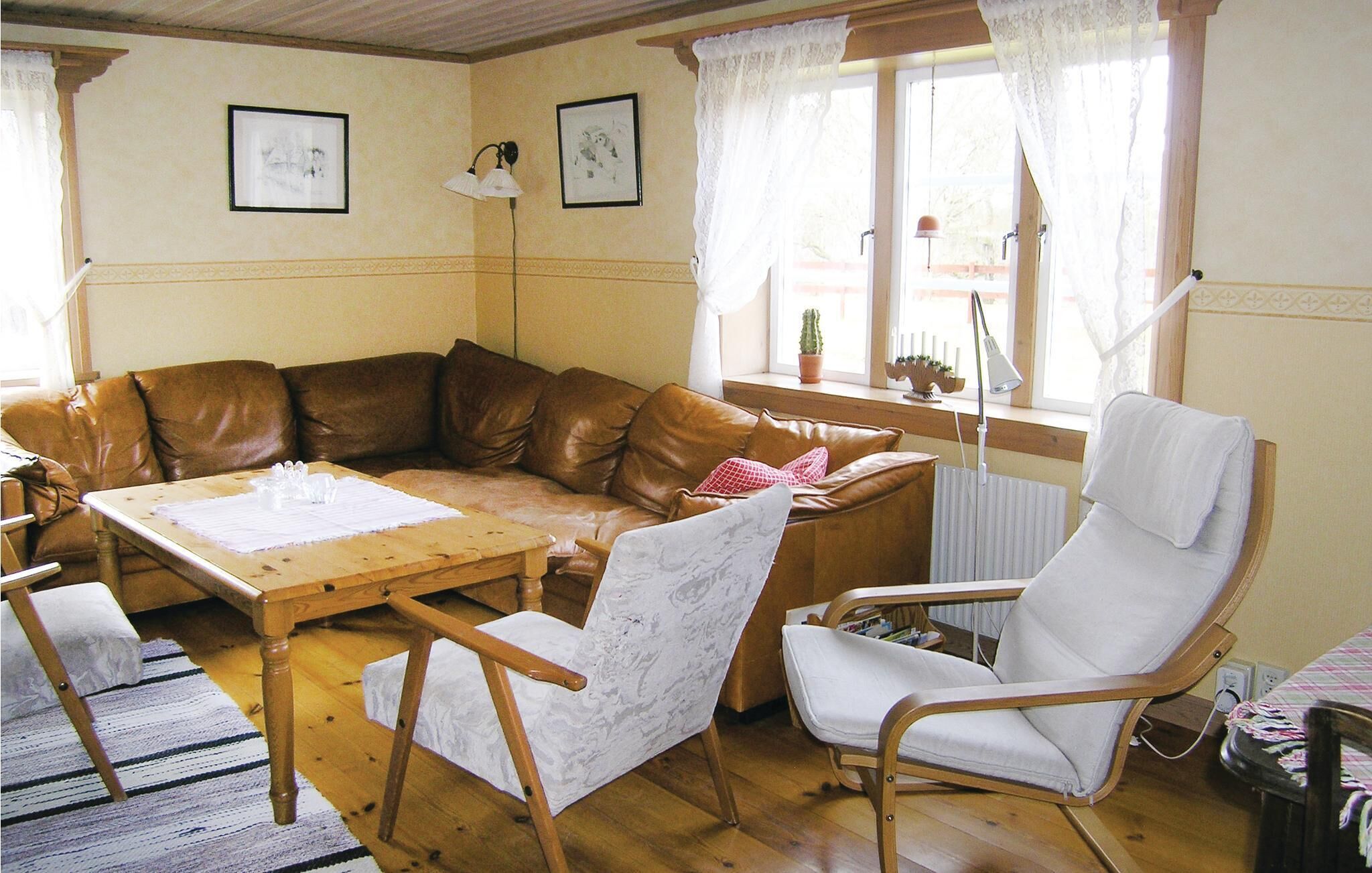 A cozy living room with a brown leather sofa, a wooden coffee table, and two white armchairs. There are framed pictures on the wall, a window with curtains, and a radiator.
