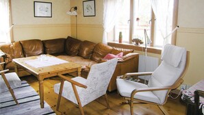 Ein gemütliches Wohnzimmer mit einem braunen Ledersofa, einem Holztisch und zwei weißen Sesseln. An der Wand hängen gerahmte Bilder, ein Fenster mit Vorhängen und ein Heizkörper.