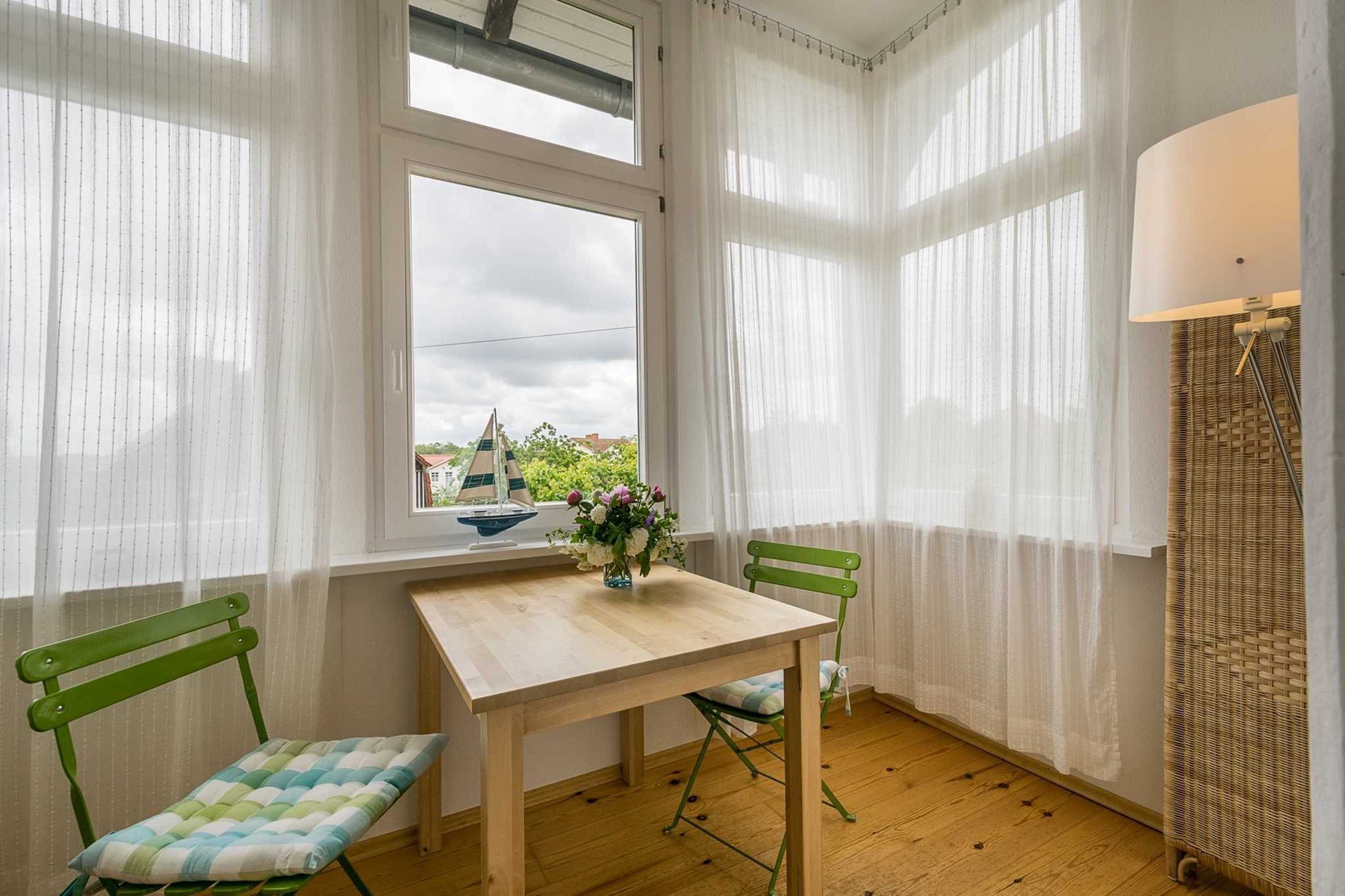 Ein Zimmer mit einem Holztisch, zwei grünen Stühlen, einem Fenster mit Blick auf Grünflächen und ein Segelboot sowie einer Lampe auf einem Korbständer.