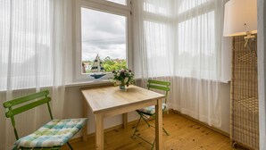 Een kamer met een houten tafel, twee groene stoelen, een raam met uitzicht op groen en een zeilboot, en een lamp op een rieten standaard.
