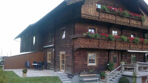 Un chalet in legno con un balcone decorato da fiori rossi, un patio con mobili da esterno e una scala che conduce all'ingresso.