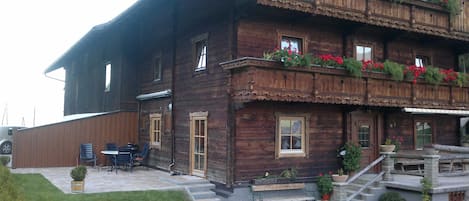 A wooden chalet with a balcony adorned with red flowers, a patio with outdoor furniture, and a staircase leading to the entrance.