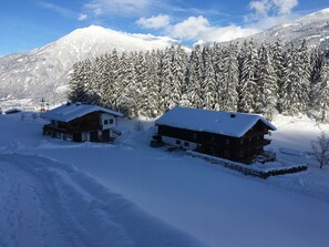 Zwei Holzhütten mit schneebedeckten Dächern in einer verschneiten Berglandschaft.