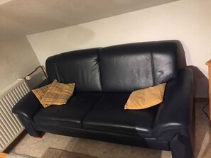 Ein schwarzes Ledersofa mit zwei karierten Kissen in einem Raum mit Heizkörper und einem Holztischchen.