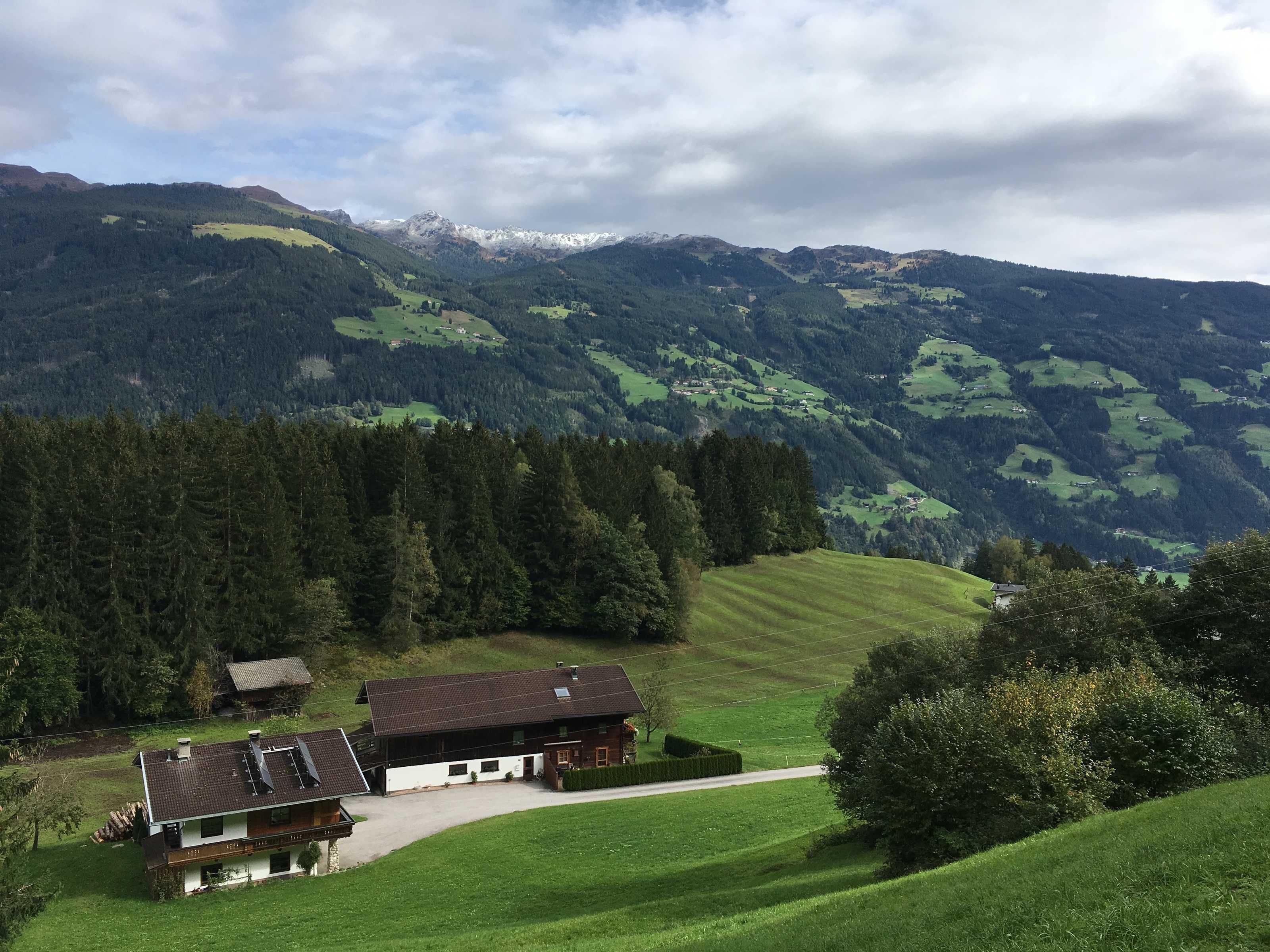 A rural house with a sloped roof, surrounded by green fields and a forested mountain range.