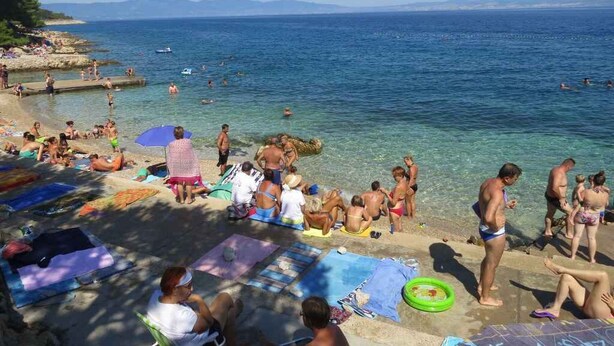 A beach with people sunbathing and swimming, clear blue water, and distant mountains.