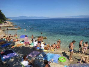 Ein Strand mit badenden Personen, klarem, blauem Wasser und Bergen im Hintergrund.