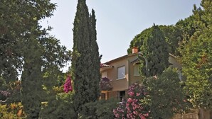 Un bâtiment à deux étages, doté d’une façade beige, entouré d’une végétation luxuriante et de grands arbres.