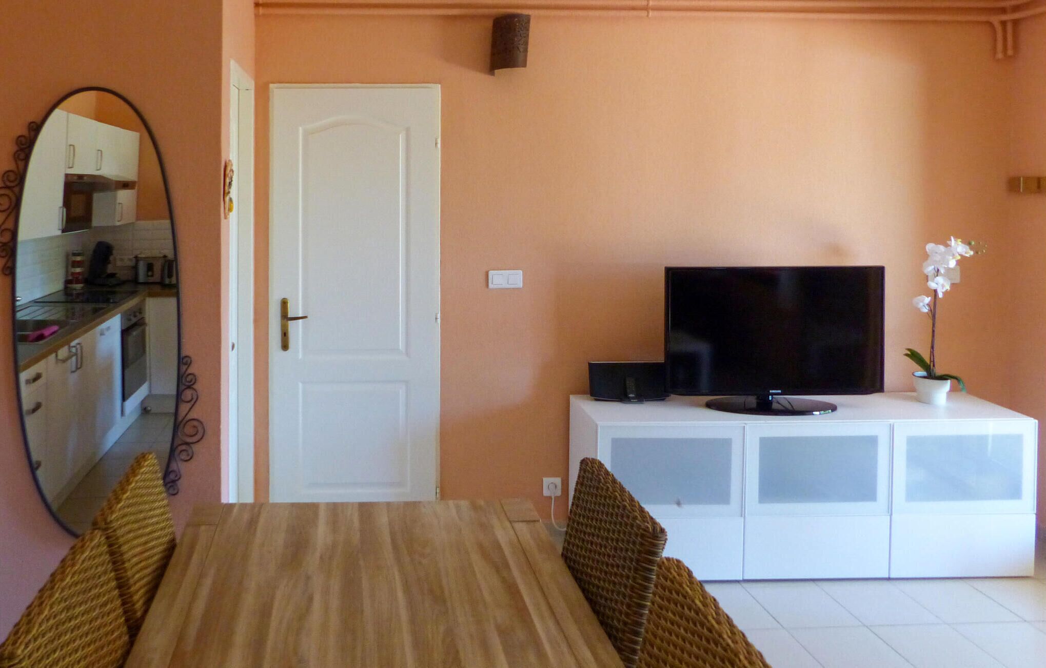Una habitación con una mesa de comedor, un televisor sobre un mueble blanco, una puerta y una cocina visible en un espejo.