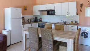 Une cuisine compacte avec des meubles blancs, un four à micro-ondes, une cuisinière et un lave-linge. On y trouve une table à manger avec des chaises en osier.