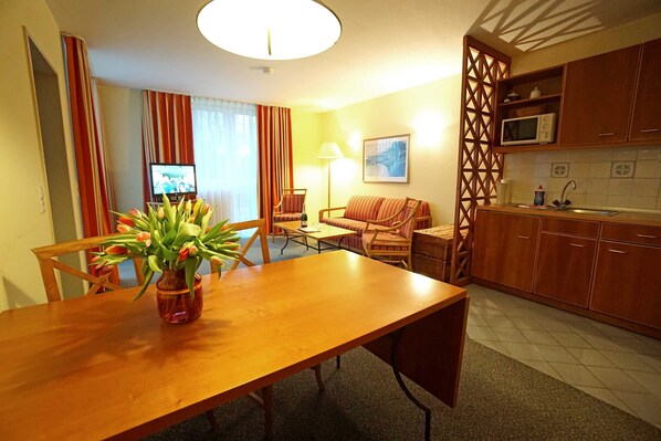 A dining area with a wooden table, chairs, a vase of flowers, a sofa, and a television in the background.