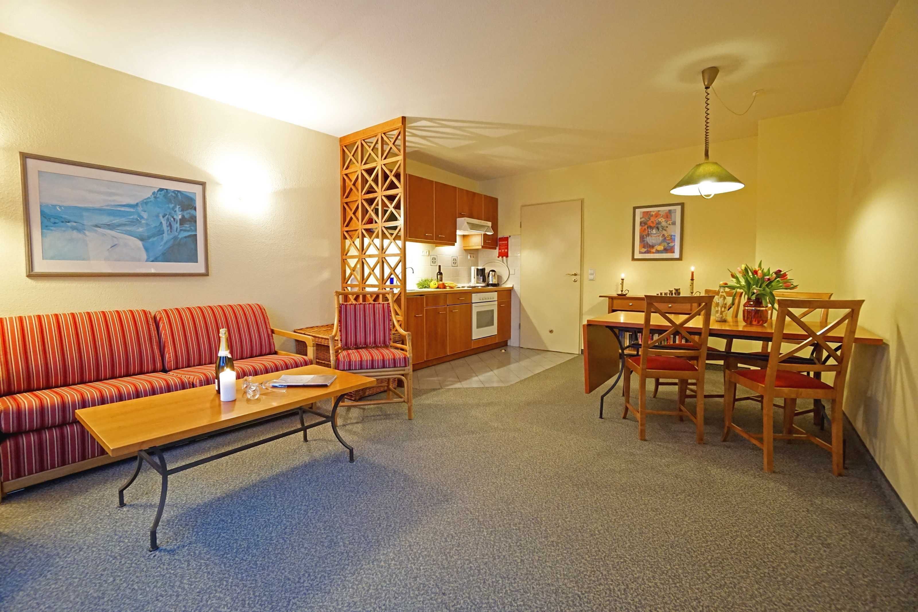 Ein Wohnzimmer mit Sofa, Couchtisch und Essbereich mit Stühlen. Im Hintergrund ist eine Küche zu sehen.