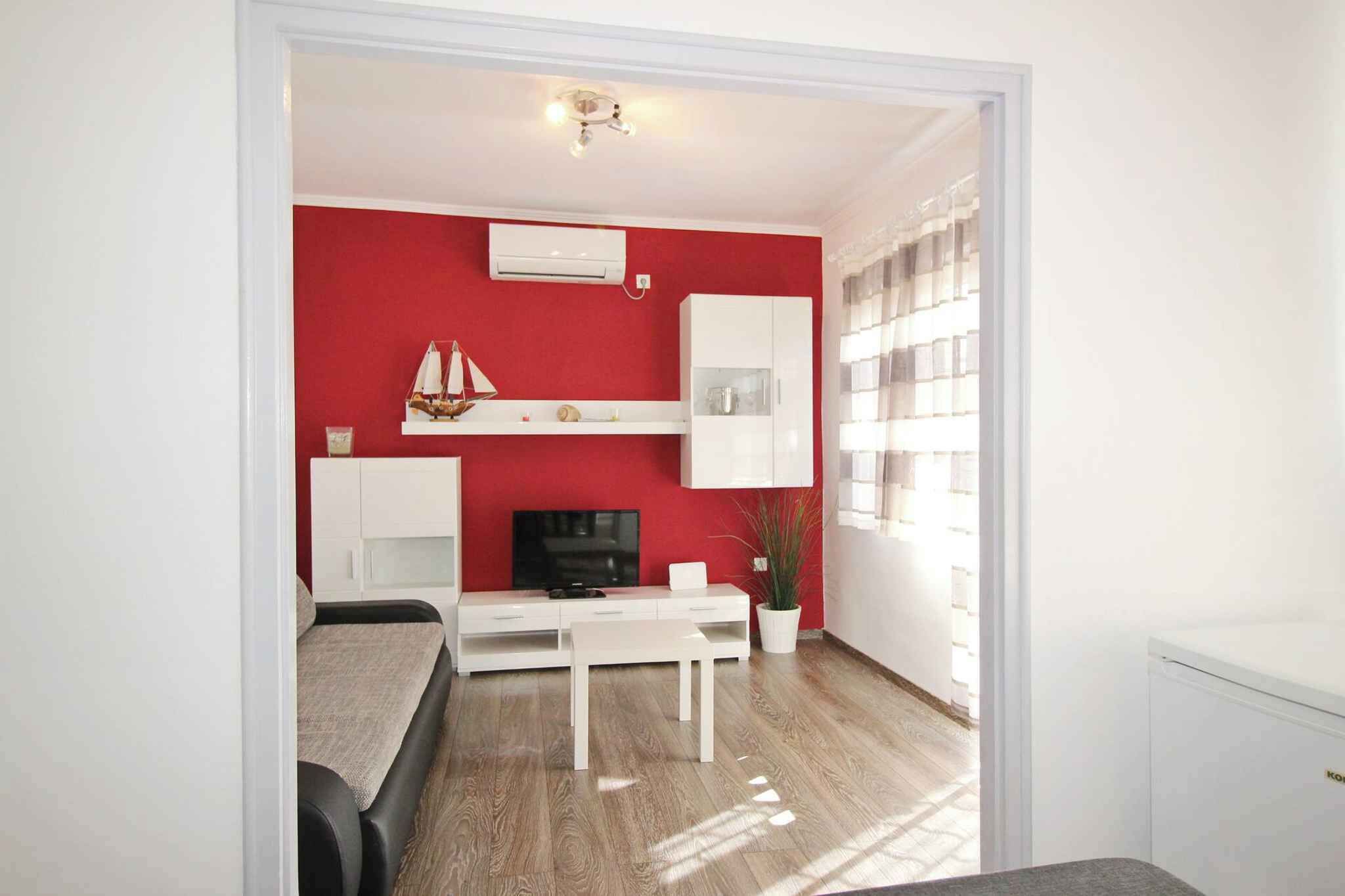 A compact living space with a red accent wall, white furniture, a small sailboat decoration, and a window with blinds.