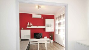 Un espacio habitable pequeño con una pared de acento roja, muebles blancos, un adorno de velero y una ventana con persianas.