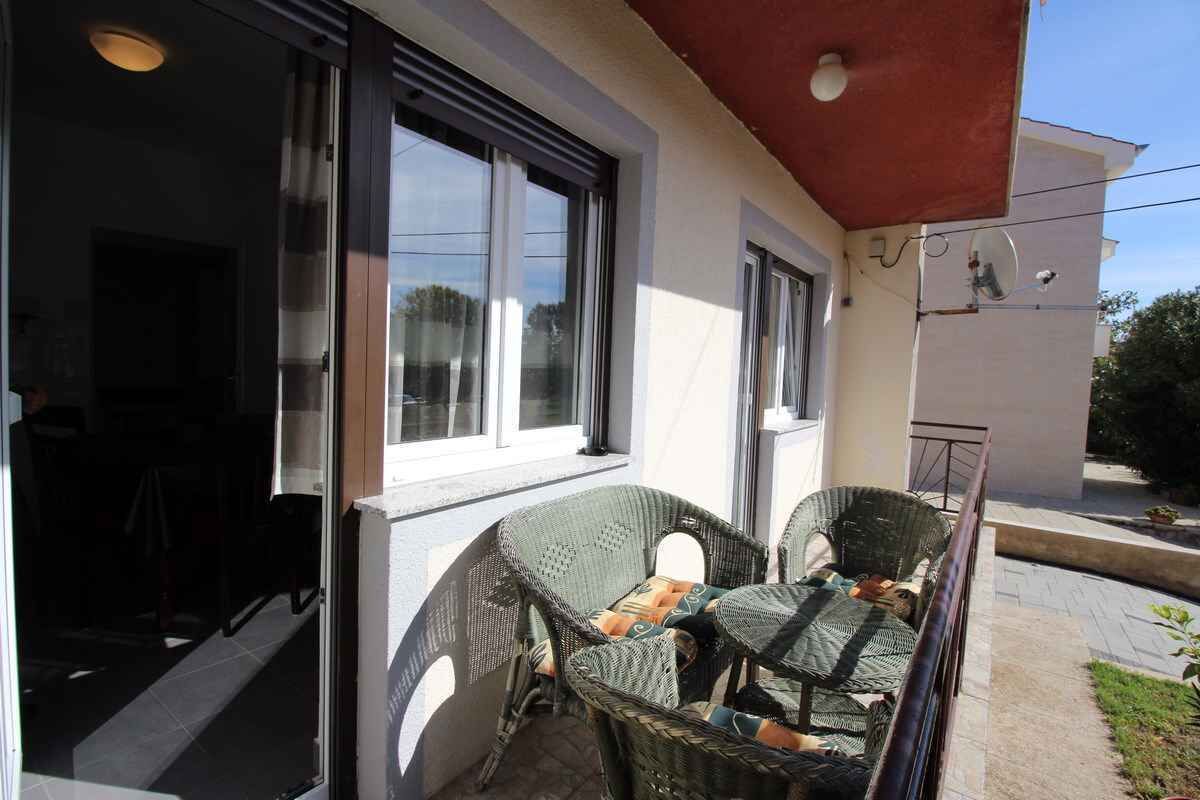 A balcony with wicker chairs and a table, overlooking a paved area with a building in the background.